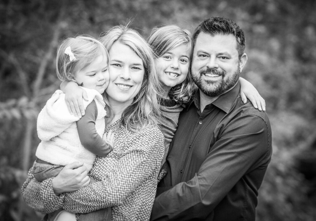 A smiling family of four posing outdoors, featuring two parents holding their young daughters, captured in a black-and-white portrait.